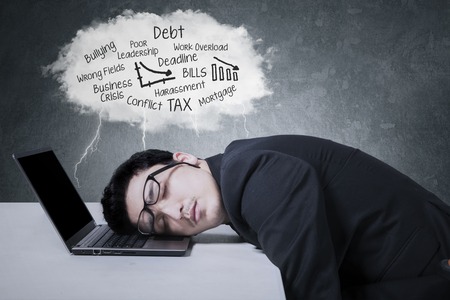 Male worker sleeping on a laptop computer under a cloud bubble of his problemsの写真素材
