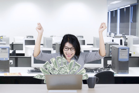 Cheerful Asian businesswoman looking at dollars cash on the laptop screen in the office roomの写真素材