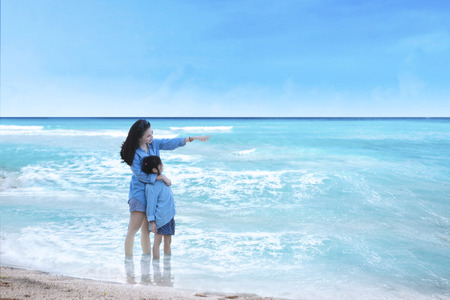 Young woman and her daughter standing on the beach while looking at the seaの写真素材