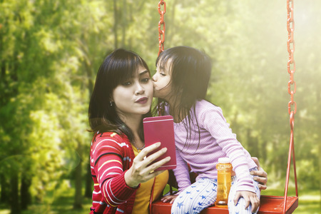 Happy young mother and her daughter taking selfie photo together while the girl kissing her mother at the parkの写真素材