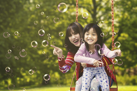 Cheerful Asian woman and little girl sitting on a swing while playing soap bubbles, shot at the parkの写真素材