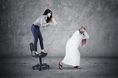 Picture of young businesswoman shouting to her arabian employee with a megaphone while standing on the chairの写真素材