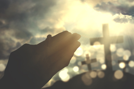 Silhouette of human hands praying to the GOD while holding a rosary with a crucifix symbol and sunbeam on the skyの写真素材