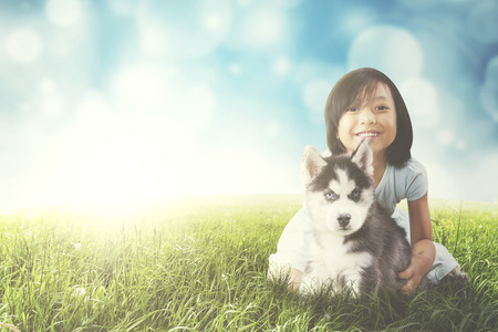 Image of a cute girl playing with a husky puppy while sitting on the grass with light glitter backgroundの写真素材