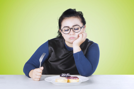 Image of overweight woman feeling bored with donuts on a plate while holding forkの写真素材