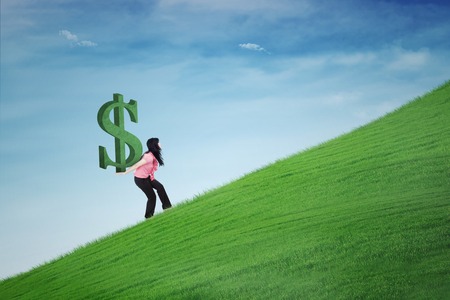 Young businesswoman carrying a dollar symbol on her back while climbing a hillの写真素材
