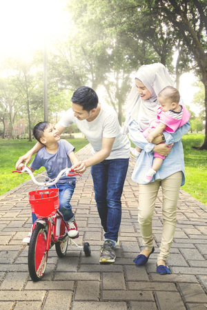 Portrait of little boy learns to ride a bicycle with his family on the park while her mother holding babyの写真素材