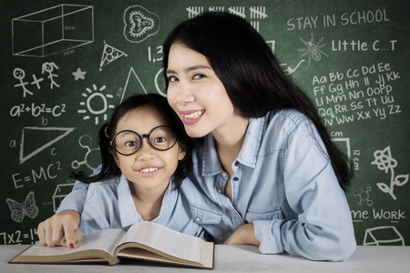 Image of female teacher with her student learning together while smiling at the camera and sitting in the classroom with doodles on the chalkboardの写真素材