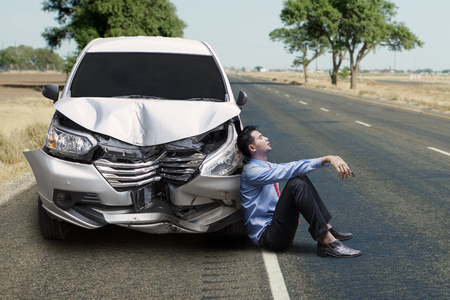 Portrait of a middle eastern businessman looks frustrated while sitting near a broken car on the roadsideの写真素材