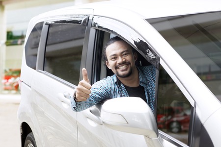 Portrait of happy Afro man showing thumbs up in his new car at the showroomの写真素材