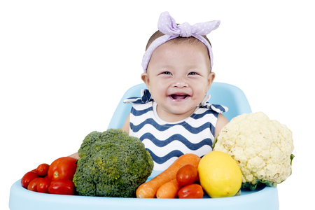 Smiling baby girl sitting on baby chair with fresh vegetables, isolated on white backgroundの写真素材
