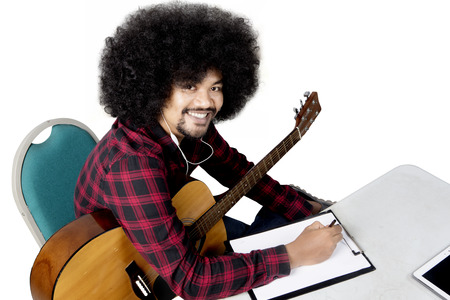Afro man listening music with earphone while writing on clipboard and holding a guitar, isolated on...
