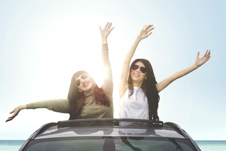 Young cheerful women wearing sunglasses, enjoying freedom on a car sunroof on the beachの写真素材