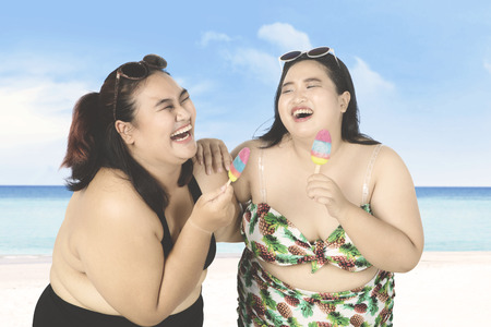 Two joyful overweight women having fun together on the beach while wearing bikini, eating ice cream, and laughing togetherの写真素材