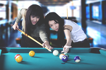 Two beautiful women playing billiard together while having fun at the bar