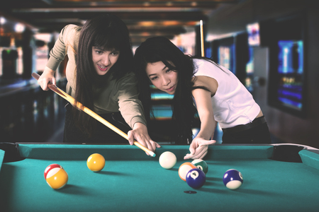 Two pretty multiracial women playing billiards together while aiming on the billiards ball, Shot at the barの写真素材