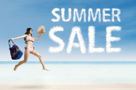 Image of pretty female enjoying a summer holiday on the beach while jumping with summer sale cloudの写真素材