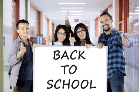 Young students showing ok sign while holding whiteboard with back to school text in the corridorの写真素材