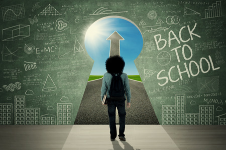 Backside of Afro student carrying backpack and walking through a keyhole with text of Back to School and upward arrowの写真素材