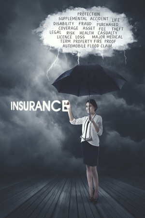 Pretty businesswoman writing a text of insurance and holding umbrella under a cloud bubble of problemsの写真素材