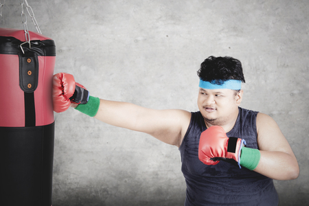 Diet concept. Portrait of a young overweight person doing exercise by punching a boxing sack while wearing sportswearの写真素材