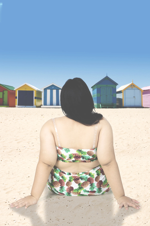 Backside of obese woman sits on seashore while wearing swimsuit and looking at colorful beach hutsの写真素材