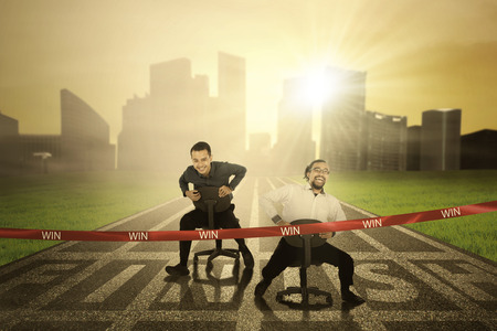 Two businessmen racing through a ribbon with word of win to the finish line while riding a chairの写真素材