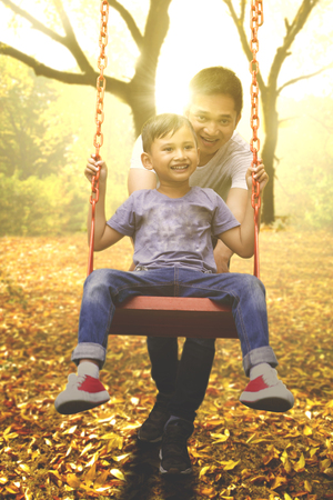 Portrait of a cheerful young father and his son playing on a swing at the park, shot at autumn timeの写真素材