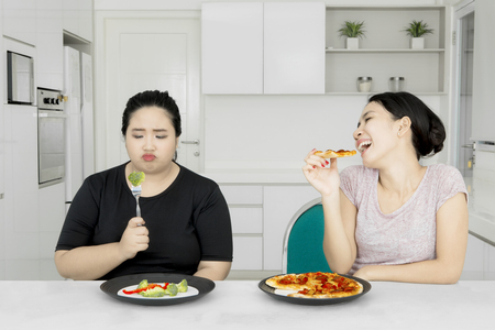 Asian woman tempting her fat sister while eating pizza and sitting together in the kitchenの写真素材
