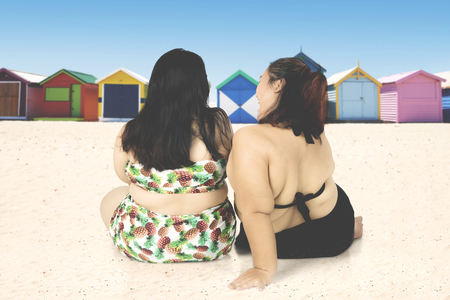 Back view of two fat women wearing swimwear while sitting on the beach sand near the cottageの写真素材