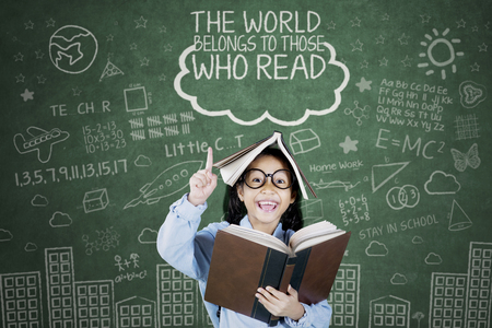 Clever schoolgirl getting inspiration with textbook over her head while standing in the classroom, motivation conceptの写真素材