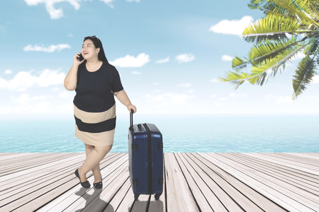 Overweight young woman standing on the wooden jetty while talking on the mobile phone and holding a suitcaseの写真素材