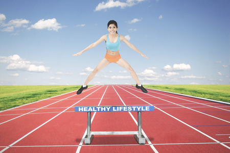 Picture of a sporty woman running on track while jumping above a text of healthy lifestyle on the hurdleの写真素材