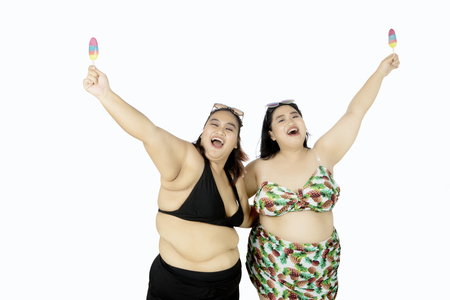 Summer concept. Two joyful overweight women having fun together while wearing bikini and eating ice cream, isolated on white backgroundの写真素材