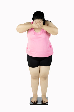 Overweight woman standing on a scale and looks worried, closing her face. Isolated on white backgroundの写真素材