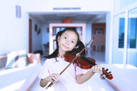 Picture of cute little girl is playing a violin while standing in the living roomの写真素材