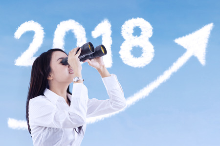 Young businesswoman using a binocular while looking at clouds shaped number 2018 and upward arrow in the skyの写真素材