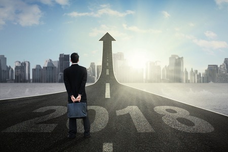 Back view of male entrepreneur holding a suitcase while looking at upward arrow and standing on the...