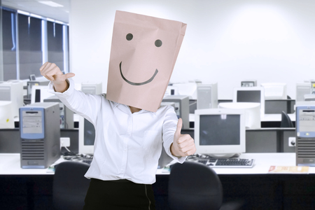 Unknown businesswoman with paper bag on her head while showing thumbs up in the workplaceの写真素材
