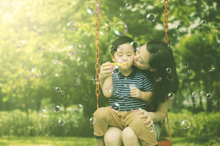Picture of young woman kissing her son while playing with soap bubbles and sitting on the swing. Shot in the parkの写真素材