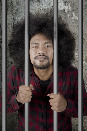 Portrait of male drunkard looking at the camera while standing behind bars. Shot in the jailの写真素材