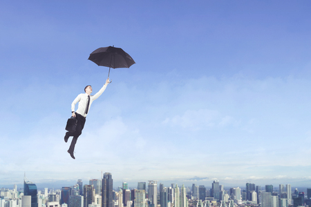 Caucasian businessman carrying a suitcase while using an umbrella to flying under blue skyの写真素材