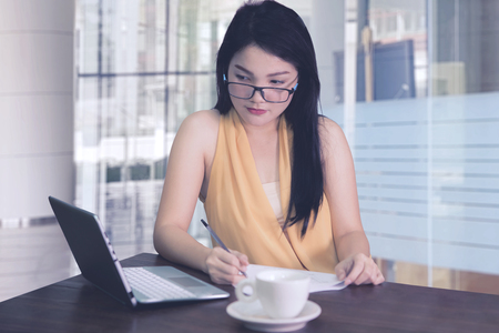 Female entrepreneur writing on the paperwork while working with a laptop in the officeの写真素材