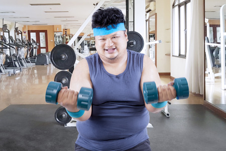 Image of obese man smiling at the camera while exercising with barbells in the gym center の写真素材