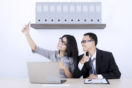 Two business people taking selfie photo while showing thumbs up in the office roomの写真素材