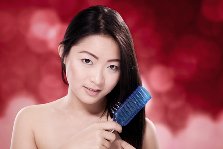 Beautiful chinese woman brushing hair against blurred red background. Asian beauty and haircare conceptの写真素材