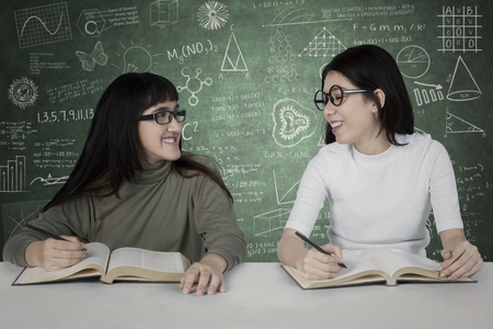 Two nerdy female students studying together while sitting in the class with scribbles on the chalkboardの写真素材