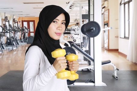 Muslim woman wearing sportswear and veil while doing workout with two dumbbells in the gym centerの写真素材