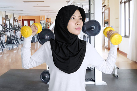 Asian woman wearing sportswear and veil while exercising with two dumbbells in the gym centerの写真素材