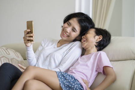 Young woman using a mobile phone while laughing with her daughter on the couchの写真素材